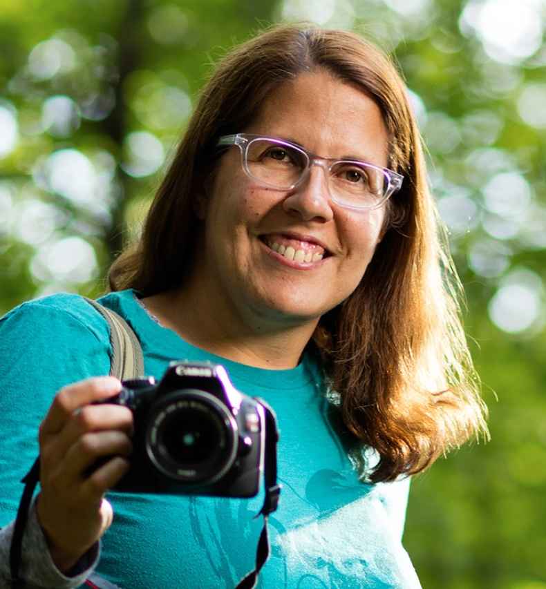 Participant Marlena - person with brown hair and glasses posing with a camera in a forest
