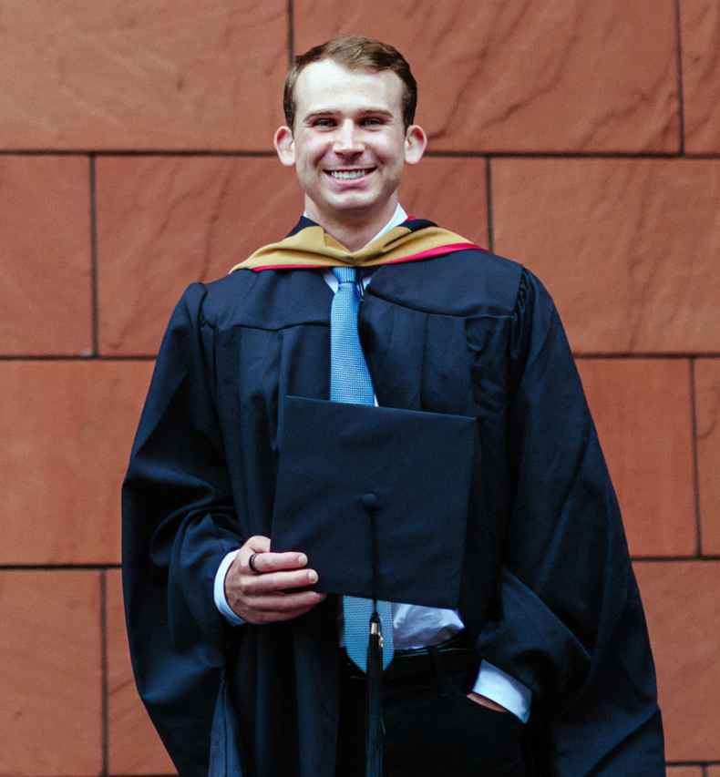 Deconstructing Stigma participant Scott - person poses in front of wall in graduation gown