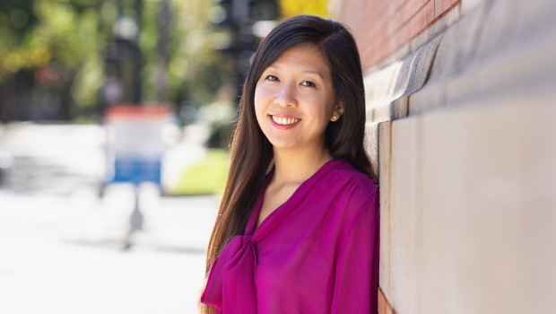 Participant Lena - person with long dark hair in a pink blouse leaning against a brick building