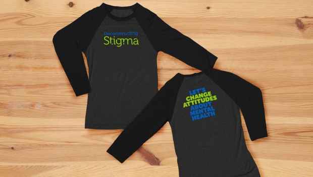 Two shirts with writing, one says Deconstructing Stigma and the other says Let’s Change Attitudes About Mental Health on a light wood background