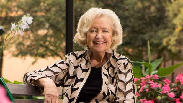 Participant Brenda - person with short white hair smiles outside among greenery and pink flowers