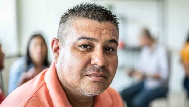 Portrait of a person with short hair wearing a coral shirt. There are other people in the background