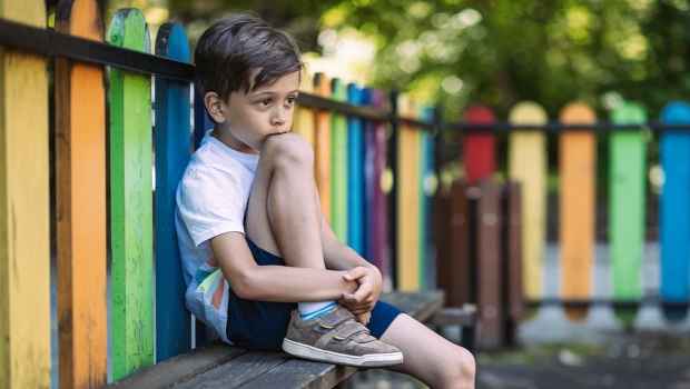 Young child sitting with a knee up on a bench in front of a colored wooded fence