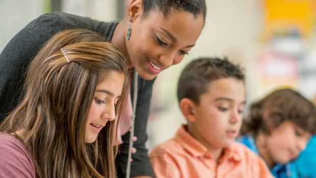 Woman in a classroom leaning over the shoulder of a student to see what is on the desk in front of them