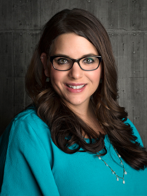 Woman with glasses, turquoise shirt and brunette hair looking at camera in front of gray background