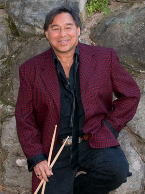 Man in front of stone wall in red blazer, holding drumsticks and smiling at camera