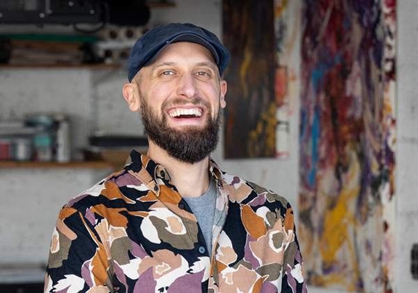 Man in hat with beard smiling in front of artwork and guitar